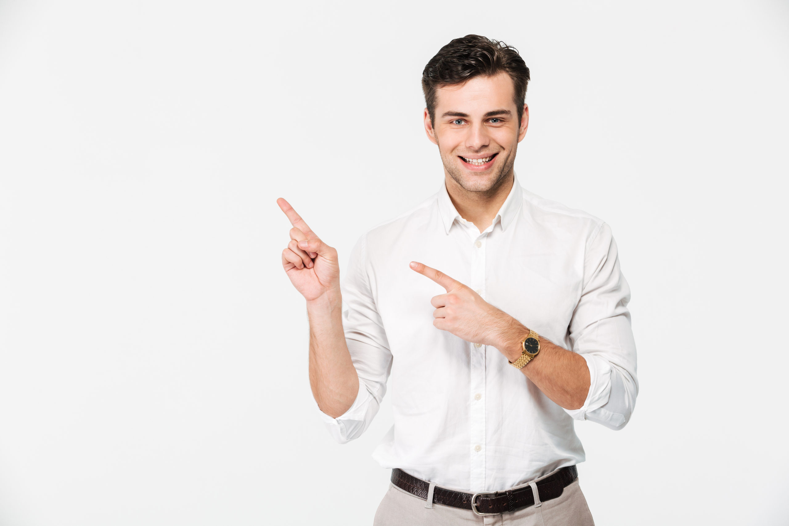 Portrait of a joyful young man in white shirt pointing two fingers away at copy space while standing and looking at camera isolated over white background
